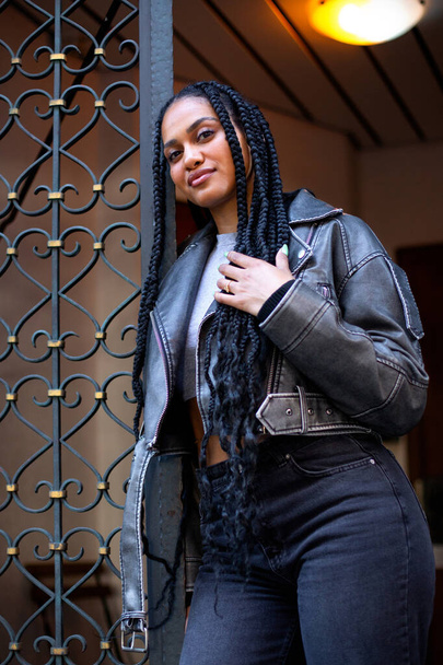 schöne junge schwarze Frau mit Afro-Zöpfen in Lederjacke hält sich in der Nähe des Eingangs auf - Foto, Bild