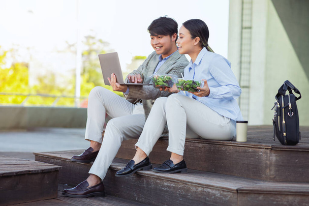 Sentados em degraus fora de um prédio de escritórios, o homem e a mulher chineses vestidos com trajes casuais de negócios estão envolvidos em uma tarefa colaborativa usando um laptop, almoçar - Foto, Imagem
