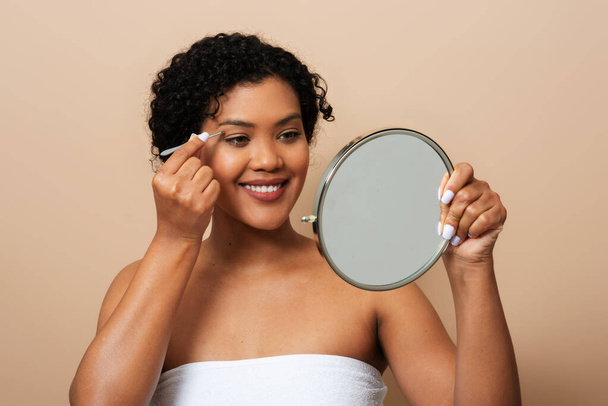 A young Hispanic woman is standing in front of a mirror, carefully applying makeup to her face. She is focused and precise, using various tools and products to enhance her features. - Photo, Image