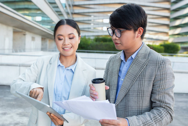 Two Asian business professionals, a man and a woman, are focused on studying a document together. Their expressions are serious and attentive as they analyze the information presented. - Photo, Image