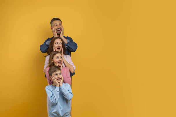 Surprised family of four, with two children and their parents, is lined up vertically, each placing their hands on their cheeks in a playful expression of surprise and happiness, copy space - Photo, Image
