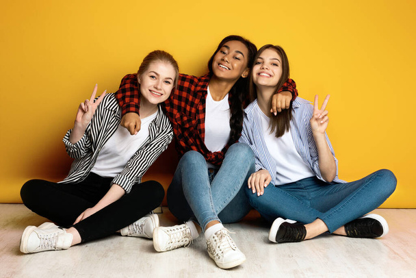 Tři mladé dívky sedí na podlaze a zaujímají různé postoje, jako by se připravovaly na skupinovou fotografii. Vypadají vesele a vzrušeně, tvoří různé pózy s rukama a nohama. - Fotografie, Obrázek