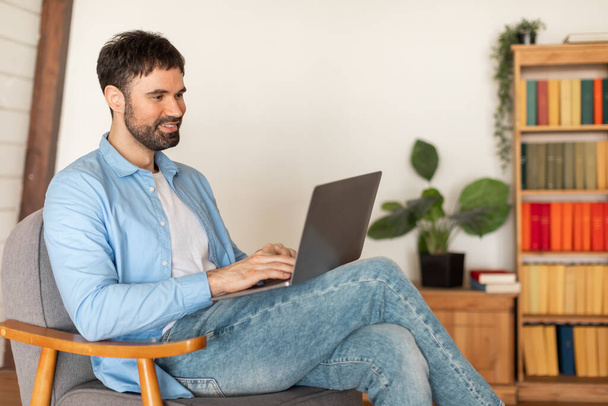 Um homem está sentado em uma cadeira, focado em uma tela de computador portátil. Suas mãos estão digitando no teclado enquanto ele trabalha. O quarto parece estar bem iluminado, espaço de cópia - Foto, Imagem