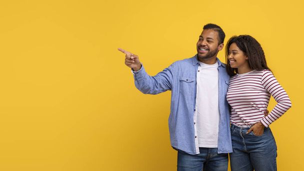 Ein afroamerikanisches Paar zeigt auf ein unsichtbares Objekt, während es vor einem leuchtend gelben Hintergrund steht. Beide wirken engagiert und fokussiert auf das, was sie andeuten, den Raum kopieren - Foto, Bild