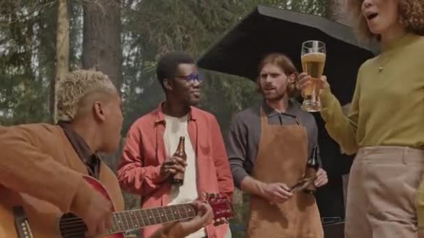 Mladý muž hraje na kytaru a zpívat spolu s přáteli stojící kolem a drží pivo a zároveň se baví na venkovní BBQ party v přírodě - Záběry, video