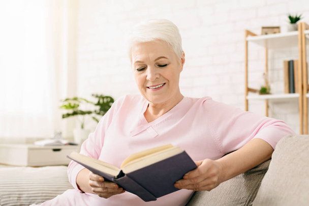 La mujer mayor está sentada en un sofá, absorta en leer un libro. Ella está enfocada en las páginas, sosteniendo el libro en sus manos. - Foto, imagen