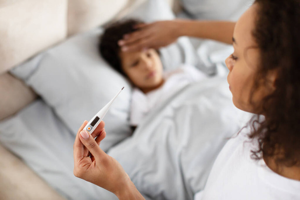 A caring African American mother is gently placing her hand on her child forehead to feel for a fever while holding a digital thermometer. The child appears to be resting in bed - Photo, Image