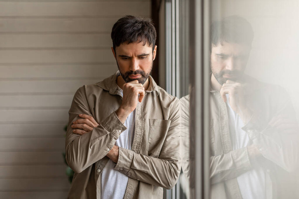 A contemplative man leaning on a large window pane, with his reflection visible. He appears deep in thought, resting his chin on his hand. His expression is one of serious contemplation or concern - Photo, Image