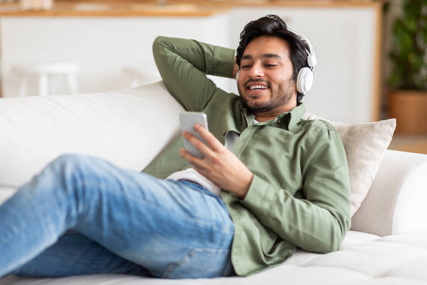 El hombre árabe está sentado en un sofá, escuchando atentamente la música que suena desde su teléfono, usando auriculares. Parece relajado mientras disfruta de la música a través de sus auriculares. - Foto, imagen