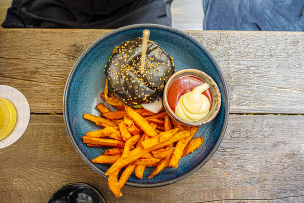 Černý burger s hranolky na dřevěném pozadí sloužil na modrém keramickém talíři - Fotografie, Obrázek