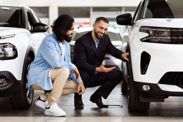 Two men are inspecting a car closely inside a well-lit showroom. One man is pointing at the features while the other is listening attentively. The car appears sleek and modern - Photo, Image