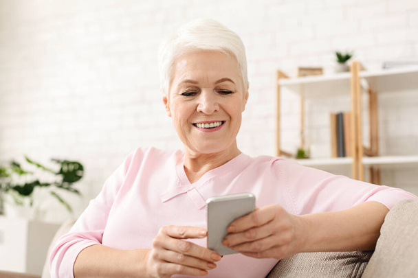 Senior woman is sitting on a couch, holding a cell phone and looking at the screen. She appears focused on the phones display, with her legs crossed and a relaxed posture. - Photo, Image