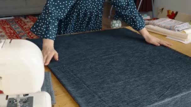 Una vista aérea de una mujer en una blusa de lunares preparando tela de mezclilla en una mesa al lado de una máquina de coser, con varios materiales de fabricación en el fondo. - Imágenes, Vídeo