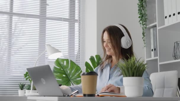 A woman works from home on the Internet. The concept of online learning and distance education. A student in headphones is working on a laptop and a cup of coffee. The freelancer, smiling, creates - Footage, Video