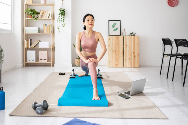 Mujer asiática joven estirando las piernas mientras ve en línea clase de fitness virtual en el ordenador portátil en casa. Concepto de fitness y estilo de vida saludable. - Foto, imagen