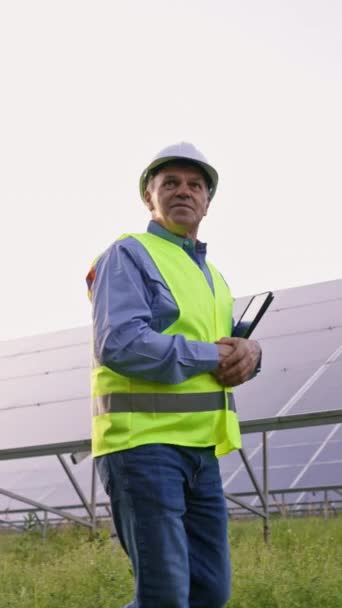 Mature engineer man in solar panels industrial area. Professional mature man technician in hardhat walks on new ecological solar construction outdoors. Concept of electricity, ecology, technologies - Felvétel, videó