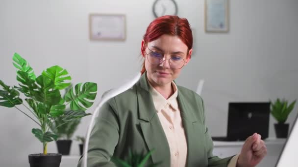 Porträt einer jungen lächelnden Frau mit Brille, die am Computer arbeitet und Notizen im Notizblock macht, während sie an einem Tisch im Büro sitzt, lächelt und in die Kamera blickt - Filmmaterial, Video
