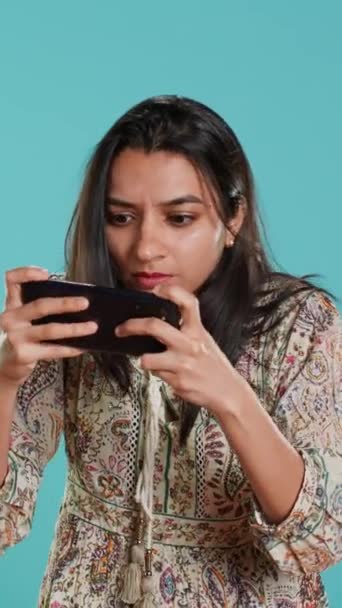 Вертикальне відео Gamer з розчарованим виглядом обличчя після отримання гри на екрані, тримає смартфон. Індійська жінка засмучена після втрати відеоігри, граючи з телефоном, ізольована на студійному фоні - Кадри, відео