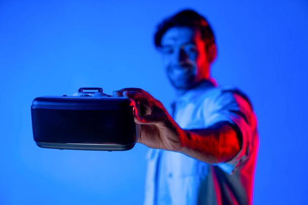 Lächelnder Kaukasier mit VR-Headset und verschwommenem Hintergrund im Stehen mit Neonlicht. Smart Teenager präsentieren digitale Brillen, um Virtual-Reality-Spiel oder Metaverse zu betreten. Abweichung. - Foto, Bild