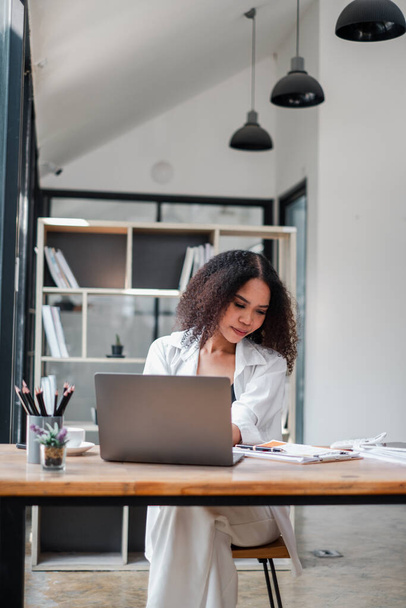 Egy nő ül egy asztalnál egy laptoppal a szeme előtt. Fehér inget és fekete nadrágot visel. A szoba modern és professzionális hangulatú, háttérben könyvespolccal - Fotó, kép