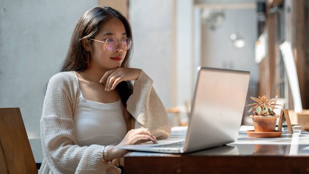 Una hermosa joven asiática con gafas y ropa casual se centra en su trabajo en su computadora portátil, leyendo documentos en línea, pensando y planeando su trabajo, sentada en una mesa al aire libre de un café. - Foto, imagen