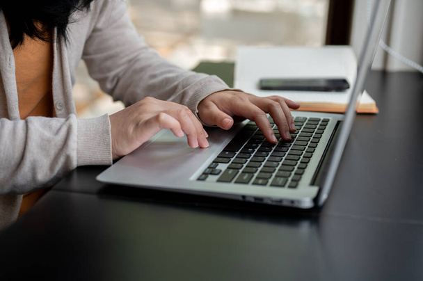 Laptop klavyesinde yazı yazan Asyalı bir kadının kırpılmış bir yan görüntüsü. Kapalı bir masada dizüstü bilgisayarı üzerinde çalışıyor. Ofiste çalışıyor ya da kahve dükkanında uzaktan çalışıyor.. - Fotoğraf, Görsel