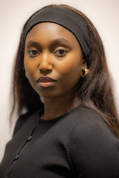Dit close-up portret vangt een jonge zwarte vrouw met een serene uitdrukking, gekleed in een klassiek zwart topje met een eenvoudige hoofdband en elegante oorbellen. Haar blik is direct en boeiend - Foto, afbeelding