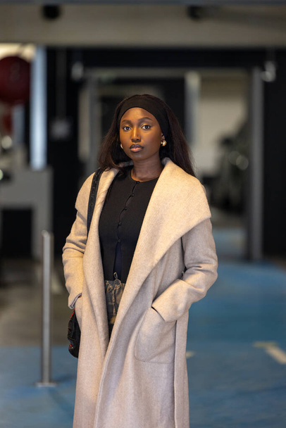 This portrait captures a young Black woman exuding sophistication and grace, dressed in a stylish light beige coat over a chic black outfit. Standing in front of an urban business entrance, her poised - Photo, image