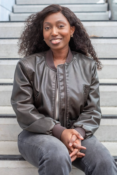 Questo vivace ritratto cattura una gioiosa donna afroamericana seduta su gradini urbani. È vestita con una giacca di pelle elegante e jeans casual, mostrando il suo sorriso radioso e la postura rilassata. Il - Foto, immagini