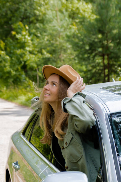 Mulher loira de chapéu a sair do carro do pára-brisas. Jovem turista explorar viagens locais fazendo momentos reais sinceros. Verdadeiras emoções expressões de fugir e refrescar relaxar ao ar livre limpo - Foto, Imagem