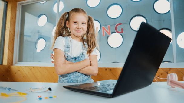 Dziewczyna za pomocą oprogramowania do programowania laptopa patrząc na kamerę z ramię złożone z pewnością świętować udany projekt. Ładny system kodowania dzieci podczas krzyżowania ramion w klasie macierzystej. Erudycja. - Zdjęcie, obraz