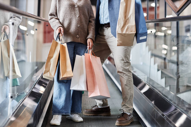 Zbliżenie nierozpoznawalnej dorosłej pary trzymającej torby na zakupy stojące na schodach ruchomych w centrum handlowym, przestrzeń do kopiowania - Zdjęcie, obraz
