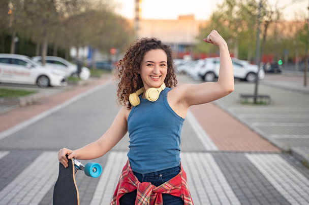 Joven mujer rizada alegre flexionando su músculo mientras sostiene un monopatín, usando auriculares y un atuendo casual en un entorno urbano al atardecer.  - Foto, Imagen