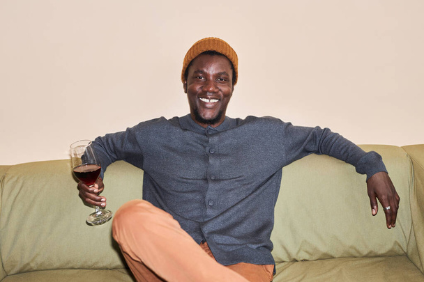 Minimal front view πορτρέτο του νεαρού μαύρου άνδρα με κόκκινο ποτήρι κρασιού lounging στον καναπέ χαμογελώντας στην κάμερα, τραβηγμένο με flash - Φωτογραφία, εικόνα