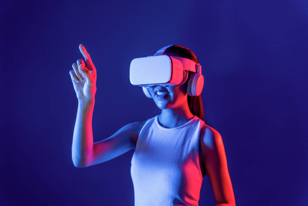Smart female stand with surrounded by cyberpunk neon light wear VR headset connecting metaverse, futuristic cyberspace community technology. Woman using finger pointing virtual object. Hallucination. - Photo, Image