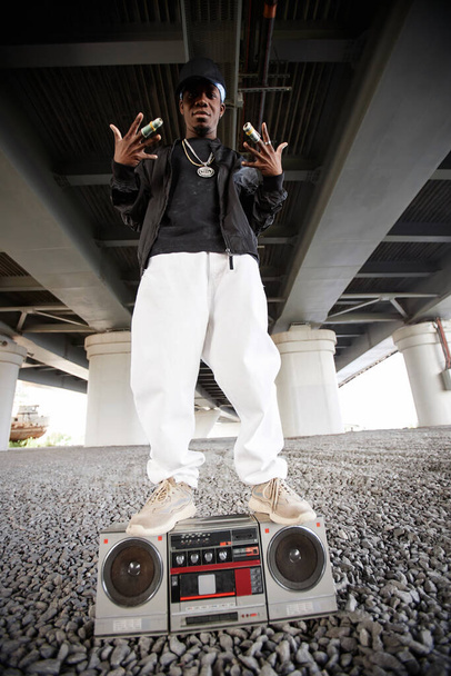 Long shot of cool rapper in trendy attire standing on top of old tape recorder in urban environment under bridge during performance - Photo, Image