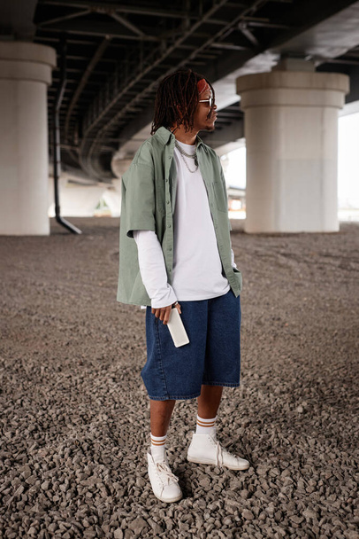 Long shot of young African American male hip hop performer in stylish casual attire standing under bridge in urban environment - Photo, Image