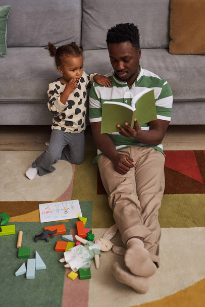Függőleges portré fiatal fekete apa olvasás könyvet a kislány ül a padlón otthon együtt - Fotó, kép
