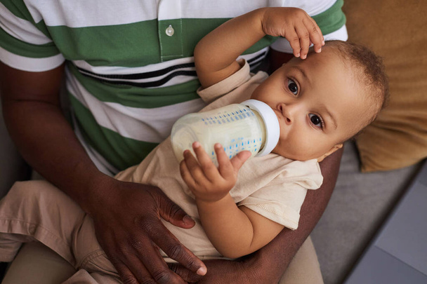 Hoge hoek portret van schattige baby jongen drinken melkfles in vader armen en kijken naar camera - Foto, afbeelding