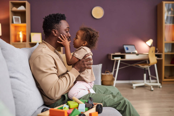 Widok z boku portret szczęśliwy Afroamerykanin człowiek przytula się z małą córką w domu siedzi na kanapie, przestrzeń kopiowania - Zdjęcie, obraz