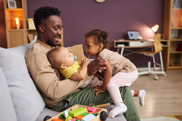 Retrato de vista lateral del feliz padre afroamericano jugando con dos niños pequeños en casa sentados en el sofá, espacio para copiar - Foto, Imagen