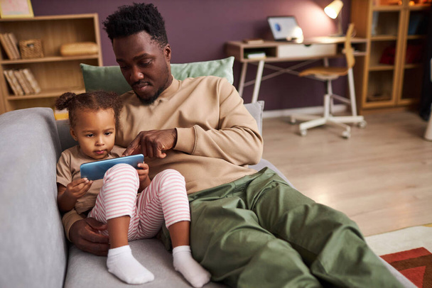 Portré fiatal fekete apa tanít kislány okostelefon használata közben együtt ülnek a kanapén otthon, másolja a helyet - Fotó, kép