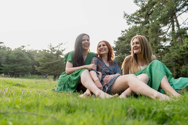 Tres mujeres disfrutan de un momento lleno de risas mientras se sientan juntas en un exuberante prado forestal, mostrando su estrecho vínculo. - Foto, imagen