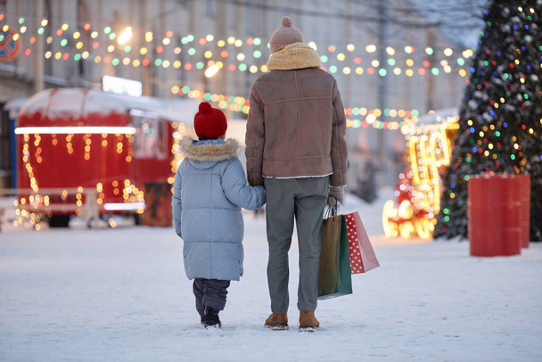Vista trasera del padre y el niño tomados de la mano caminando en el parque de invierno decorado para Navidad, espacio de copia - Foto, Imagen