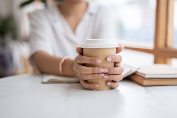 Una imagen de cerca de una mujer asiática con una camisa blanca sosteniendo una taza de café para llevar, sentada en una cafetería. bebidas, bebidas, pausa para el café - Foto, imagen