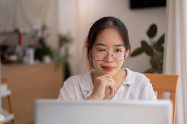 Nuori aasialainen silmälasipäinen naisopiskelija katselee kannettavan tietokoneen näyttöä, miettii ja miettii työtään, työskentelee etätyönä kahvilassa.. - Valokuva, kuva