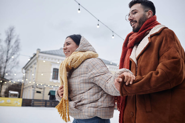 Vue latérale du jeune couple heureux patinant ensemble à l'extérieur en hiver et souriant, espace de copie - Photo, image
