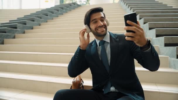 スマートフォンで笑顔でセルフィーを取る幸せなスマートビジネスマン. スーツとヘッドフォンを着用しながら階段に座っているプロのエグゼクティブマネージャーのクローズアップ画像. クリエイティブビジネス。 エクササイズ. - 映像、動画