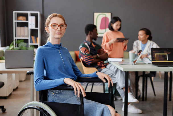 Tekerlekli sandalye kullanan genç bir kadının portresi modern ofiste kameraya bakıyor. Arka planda ekip, kopyalama alanı var. - Fotoğraf, Görsel