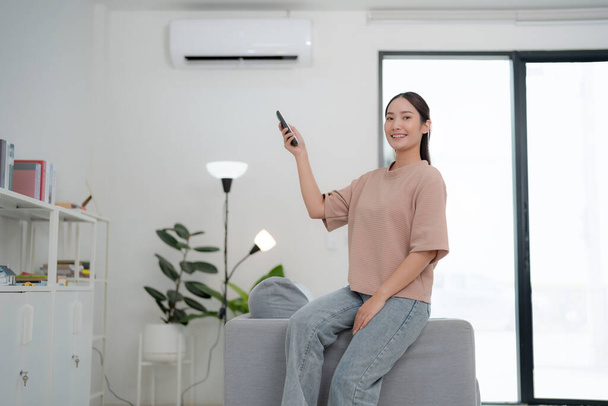 Young woman comfortably seated on a couch is using a remote control to adjust the air conditioner in a bright, modern living room with minimalist decor - Photo, Image